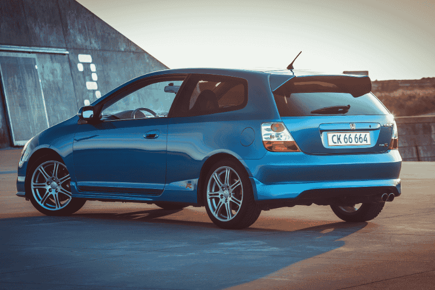 Carro hatch azul metálico com aerofólio em área pavimentada ao entardecer.