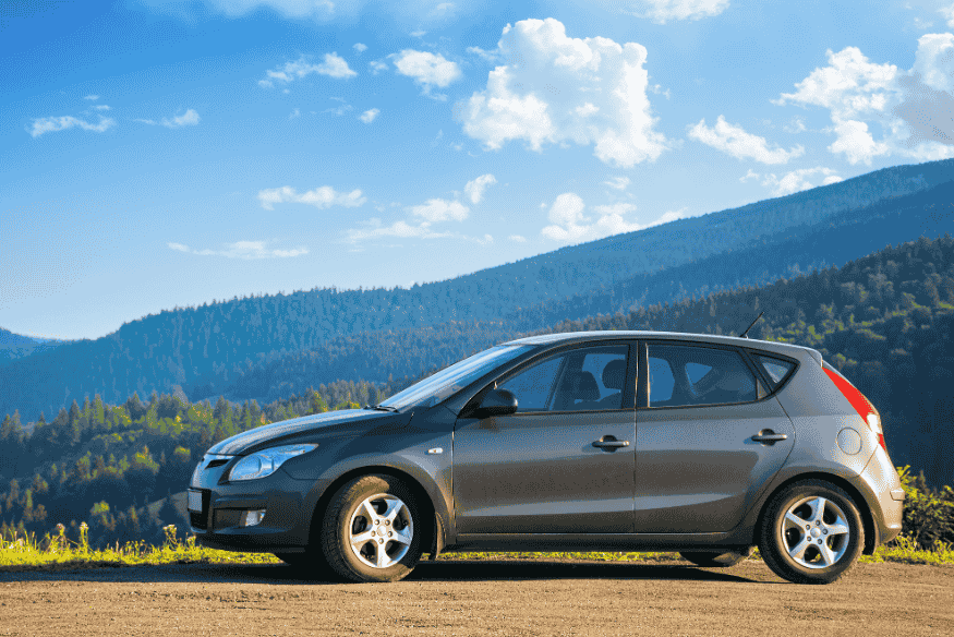 Carro hatch compacto cinza em estrada de terra com montanhas ao fundo e céu azul.