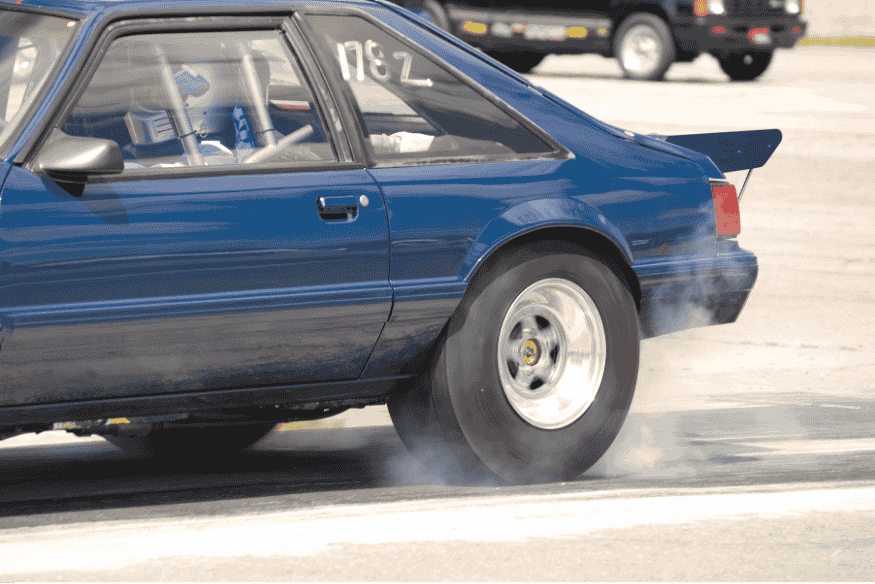 Carro de arrancada azul com gaiola de proteção e pneus traseiros gerando fumaça durante largada.