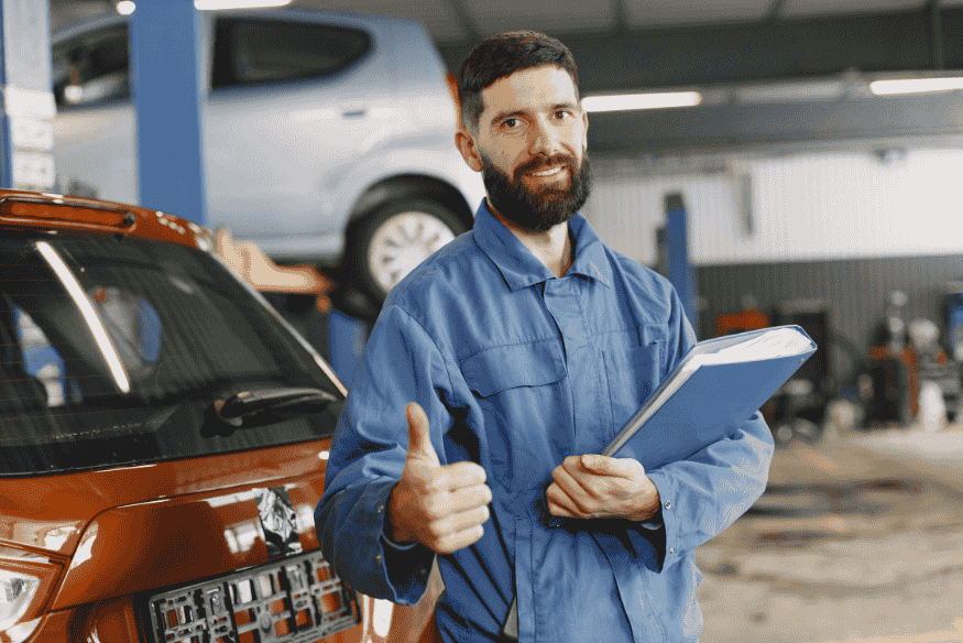 Mecânico com uniforme azul e prancheta dando sinal de positivo em oficina automotiva.