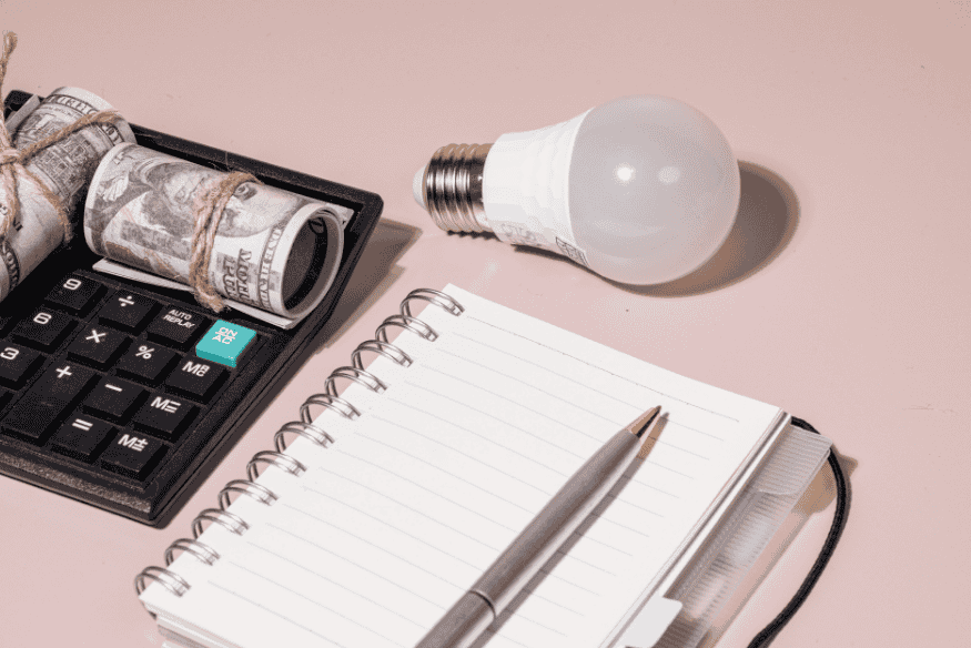 Calcolatrice con banconote arrotolate, lampadina e taccuino che simboleggiano il calcolo dei costi e le idee per ottimizzare la spesa.