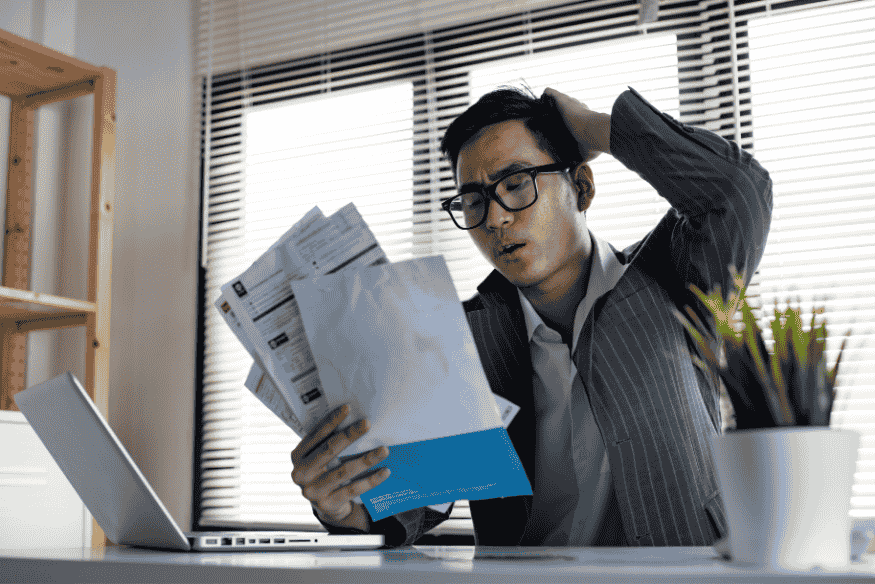 Uomo stressato che controlla documenti e fatture cartacee davanti al laptop.