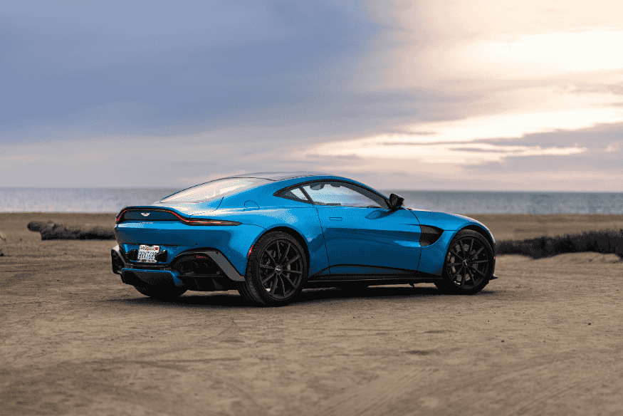 Carro esportivo azul metálico estacionado próximo ao mar, com céu parcialmente nublado.