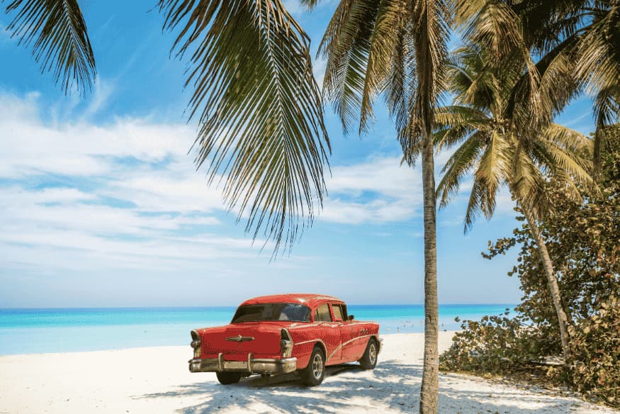 Carro antigo vermelho estacionado na areia, sob coqueiros, em frente ao mar azul.