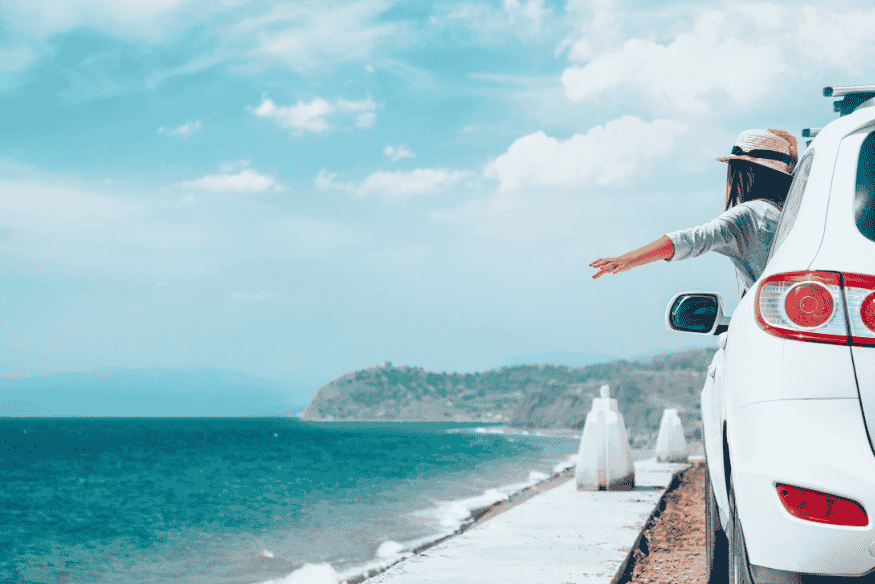 Mulher com chapéu estende o braço para fora de carro branco à beira-mar em estrada costeira.