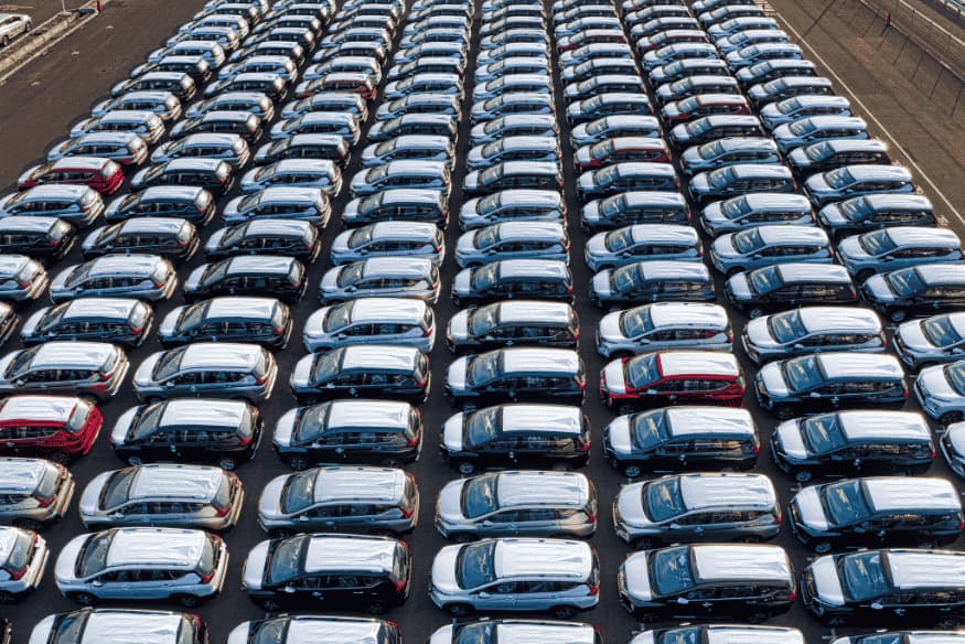Vista aérea de centenas de carros alinhados em pátio industrial, prontos para distribuição no mercado automotivo.