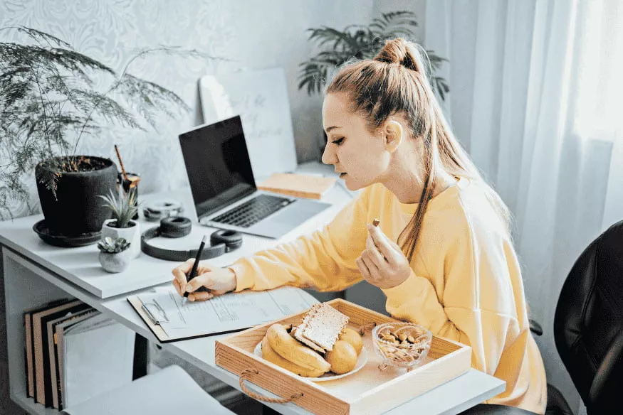 Donna che compila un modulo fiscale mentre lavora da casa con laptop e snack sulla scrivania.
