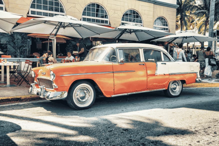 Carro clássico estacionado em rua urbana movimentada, próximo a restaurante com mesas externas