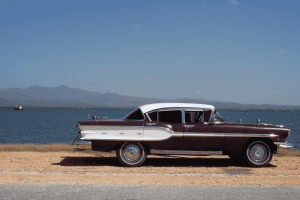Carro clássico estacionado em estrada de terra ao lado de um lago, com montanhas ao fundo