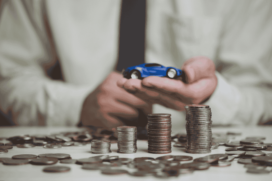 Pessoa segurando miniatura de carro com pilhas de moedas sobre a mesa, ilustrando decisão financeira