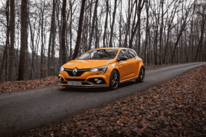 Carro hatch esportivo laranja em estrada de outono cercada por árvores secas.