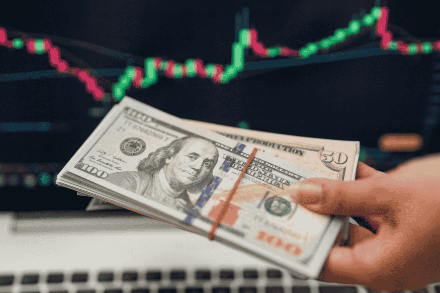 Mão com notas de dólar em frente a gráfico de variação cambial, ilustrando a relação entre câmbio e consumo