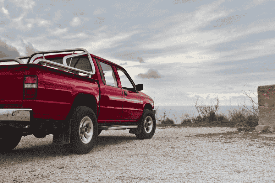 Picape vermelha clássica estacionada em terreno de cascalho com vista para o mar