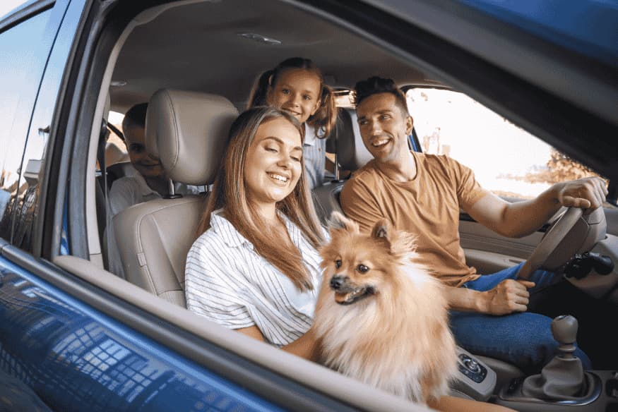 Família sorridente dentro do carro com cachorro no colo, pronta para iniciar uma viagem.